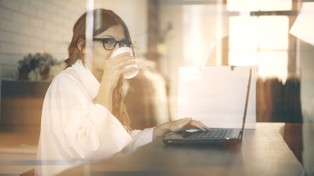 Young blonde businesswoman in glasses typing and drinking coffee in office. A side view. Toned image...
