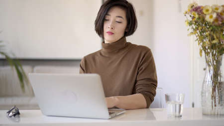 Pan shot right to left of smiling asian woman in eyeglasses working with laptop. Glass of water on table and a vase with flowers in white interior home officeの写真素材
