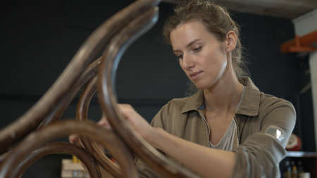 A working female carpenter peels off paint from rocking chair legs with a spatula, restorer in a workshop. Restoration of wooden rocking chair in a workshop.の写真素材