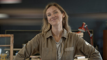 Female restorer smiling at the workshop, standing next to her working table. Young professional in furniture restoration. Portrait. Lense flareの写真素材