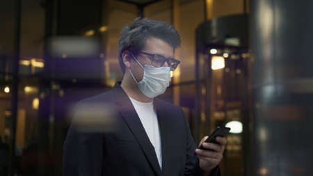 Man in mask with a phone walking on background of city glass buildings. Windy weather, man in eyeglasses and black suit scrolling the phone while walkingの写真素材