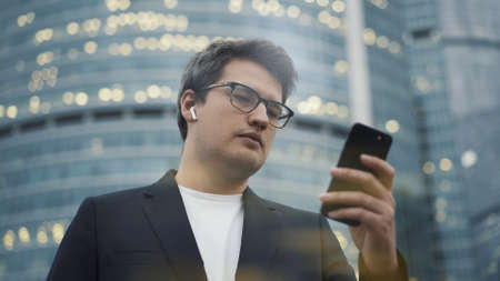 Man with a phone on background of business centre. Man dressed in black suit and white shirt scrolling the phone in earbuds, on background of glass buildings blurry effectの写真素材