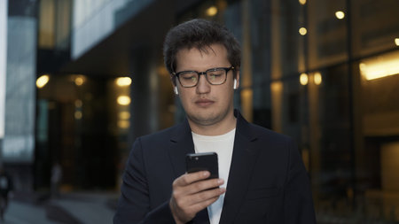 Man in earbuds scrolling the phone in evening standing on background of glass windows. Portrait shot...