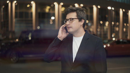 Businessman in eyeglasses calling on the phone on background of building with yellow lights, night...