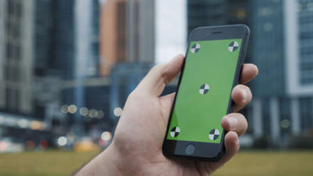 Mans hand and a phone with a green screen chroma key, on the background of a blurred buildings. Close up of man with a phone with a green screenの写真素材