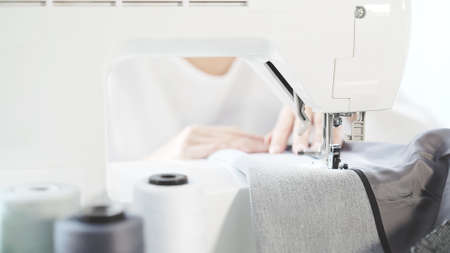 Female hands sew on a white sewing machine close-up. Concept of sewing in modern bright studio, sewing in process, blurred spool of thread on foregroundの写真素材