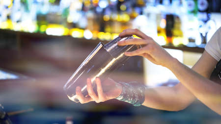 Bartender holding two metal cups for cocktail making, background of blurred bottles, lens flare. Bartender hands with two metal mixing glass for cocktailの写真素材