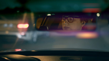 Drivers face in eyeglasses is reflected in a rear view mirror, night shot lens flare. Driver in a...