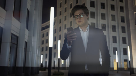 Man with phone on background of city corporate buildings, lens flare. Night shot of man in black suit and shirt wearing eyeglasses looking at the phone, late night workの写真素材