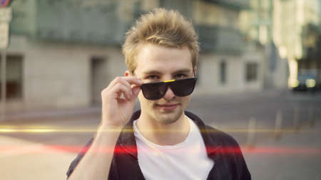Young man looking out from glasses on background of city street. Portrait shot of confident blond young man looking at the camera flirting, holding big eyeglasses lens flareの写真素材