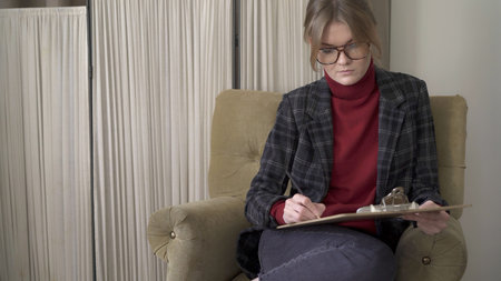Female manager in eyeglasses sitting in chair, middle shot, making notes looking at the papers. Woman in red sweater and jacket wearing big eyeglasses working in a soft chairの写真素材