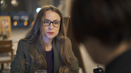 Young woman in eyeglasses with a cup of red beer sitting to table with her friend. Two women discussing in an open space bar, back of a brunette womanの写真素材