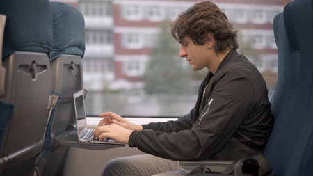 Young guy with laptop sitting in train, student in black jacket studying while traveling. Young man concentrated typing in a train, working while travelingの写真素材