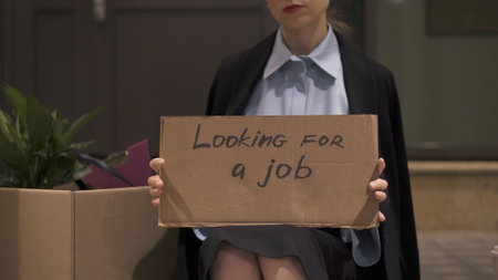 Young woman in office clothes with a sign looking for a job sitting with cardboard box. Woman in office skirt looking for a job on the street, no faceの写真素材