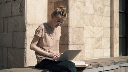 Young woman learning outside with laptop near university building, alone young student in sunglasses...