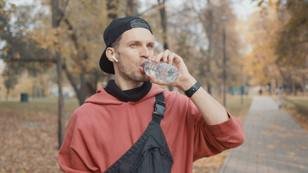 Man runner closeup in red hoodie and earbuds drinking water in the park slow motion, on background...