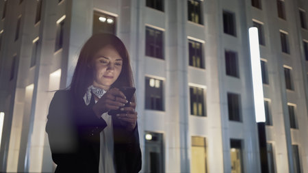Woman at night with phone wearing black jacket and neck scarf, on background of blue buildings. Middle shot of young woman alone texting in the phone, night shotの写真素材