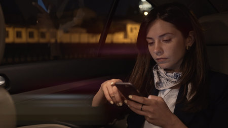 Young woman sitting in car backseat with phone in hands. Woman passenger in black jacket and neck...