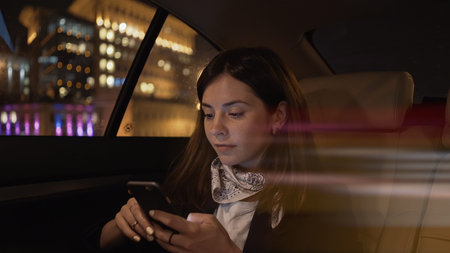 Young woman smiling sitting in car backseat with phone in hands. Woman passenger in black jacket and neck scarf alone with phone, in car late at nightの写真素材