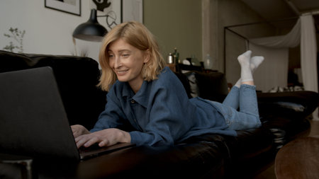 Young woman using laptop, chilling on a sofa in living room. Woman in jeans clothes concentrated, texting on a device. Concept of free time and work from home.の写真素材