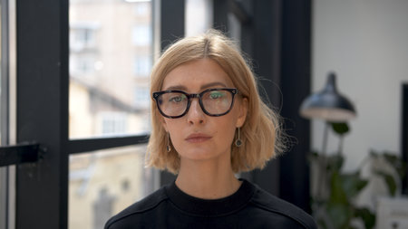 Blonde woman portrait in eyeglasses with tense emotions on background of office room. Attractive stylish person looking at the camera with neutral expression. Concept of businesswoman.の写真素材