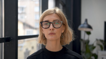 Blonde woman portrait in eyeglasses with tense emotions on background of office room. Attractive stylish person looking at the camera with neutral expression. Concept of businesswoman.の写真素材