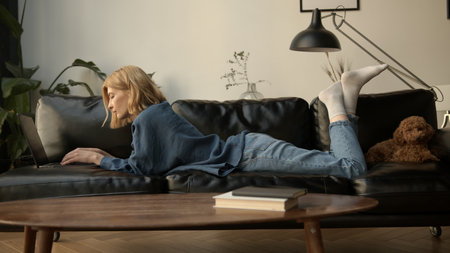 Young woman using laptop, chilling on a sofa in living room. Woman in jeans clothes concentrated, texting on a device. Concept of free time and work from home.の写真素材