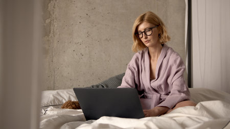 Concentrated attractive woman working with laptop on bed in bedroom in the morning. Freelancer worker during covid-19. Concept of distance work and staying home in quarantine.の写真素材