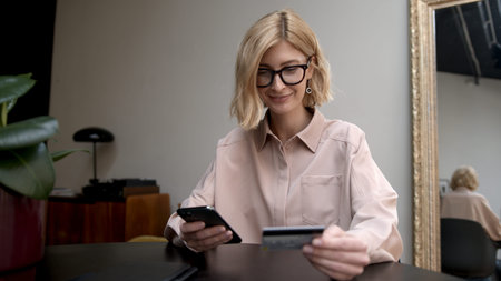 Blonde woman in eyeglasses concentrated look, phone and credit card in hands. Shopping online from home, safe payment and mobile device.の写真素材