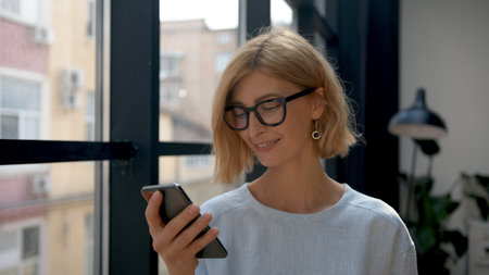 Woman holding smartphone, reading a message. Attractive blonde manager portrait on background of windows. Office room and worker with mobile phone. Concept of business and technologyの写真素材