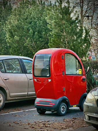 A red retro mobility scooter with a rounded roof is parked next to a silver car, set against a backdrop of trees and bushes, blending modern and vintage elements seamlessly.の写真素材