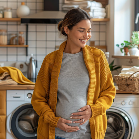A young pregnant woman in a yellow cardigan smiles in a cozy laundry room, looking at her belly. Sunlight filters through the window, creating a warm and peaceful scene.の素材