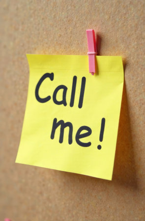 A bright yellow sticky note with bold black text invites someone to call. It is securely pinned to a corkboard, conveying a sense of urgency and communication in a simple yet effective way..の素材
