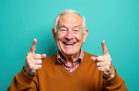 Happy elderly man with gray hair is smiling widely and pointing his fingers in excitement against a vibrant teal background, showcasing a cheerful and lively atmosphereの素材