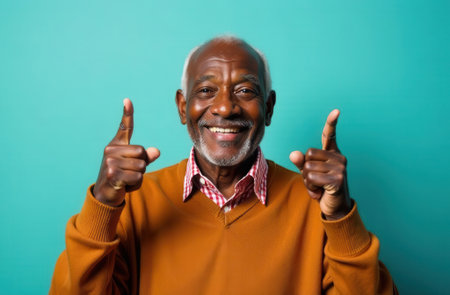 Joyful senior man with gray hair and beard is smiling widely while giving thumbs up, showcasing a positive attitude and vibrant energy against a bright turquoise backgroundの素材