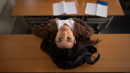 Young woman with long dark hair is lying on her desk in a classroom, surrounded by notebooks and a backpack, capturing a moment of relaxation and contemplation in an academic environmentの素材
