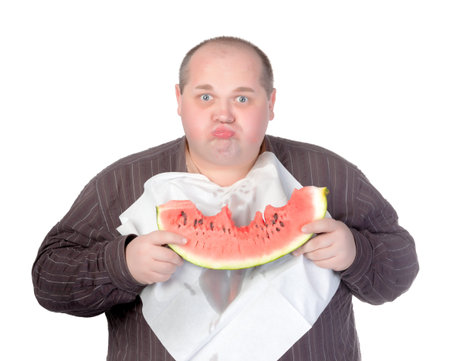 Obese man with a serviette bib around his neck standing eating a large slice of fresh juicy watermelon isolated on whiteの写真素材