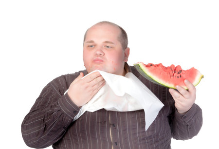 Obese man with a serviette bib around his neck standing eating a large slice of fresh juicy watermelon isolated on whiteの写真素材