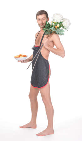 Fun image of an implied nude man clad only in a short apron bearing Valentines or anniversary gifts of a bowl of freshly baked cookies and a bunch of long stemmed flowers isolated on whiteの写真素材