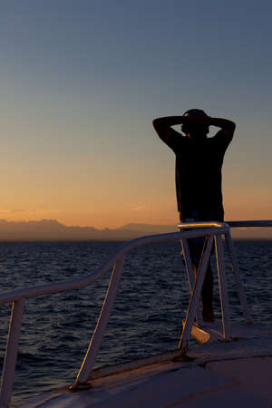 Person enjoying an ocean sunset standing on the prow of a ship looking towards the distant land and setting sunの写真素材