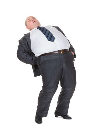 Very overweight man in a stylish suit and tie with acute back ache bending over backwards to alleviate the pain with an agonised expression on his face isolated on whiteの写真素材