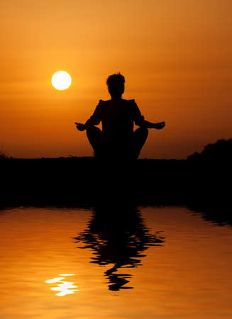 Silhouette woman sitting and relaxing against orange sunset with reflection in waterの写真素材