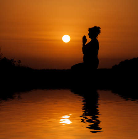 Silhouette woman sitting and relaxing against orange sunset with reflection in waterの写真素材