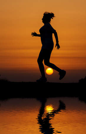 Silhouette woman running against orange sunset with reflection in waterの写真素材