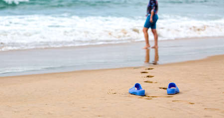 Beach Slippers in the Foreground and Blurred Silhouette of a Woman in Wavesの写真素材
