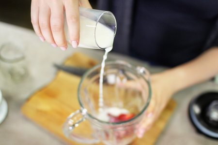 A female hand pours milk into a blender to prepare a tasty and healthy smoothie of strawberries and almond.の写真素材