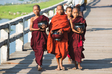 AMARAPURA, MYANMAR, JANUARY 10, 2015: Three Boys Buddhist monks walking on U Bein Bridge in traditional clothes with jug for the offerings in the morning.のeditorial素材