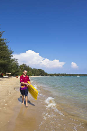 The  European man walking and carrying a yellow rubber mattress at the beach at Koh Mak island, Trat, Thailandの写真素材
