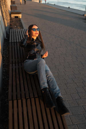 Young woman listens to music in closed headphones through her phone wearing a leather jacket and...