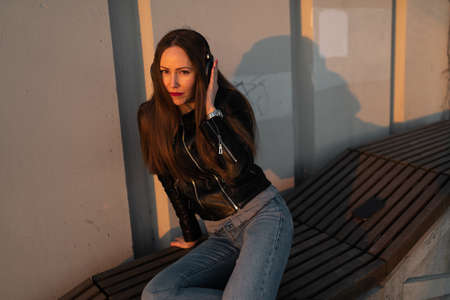 Young woman listens to music in closed headphones through her phone wearing a leather jacket and...